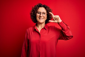 Fototapeta na wymiar Middle age beautiful curly hair woman wearing casual shirt and glasses over red background smiling and confident gesturing with hand doing small size sign with fingers looking and the camera. Measure