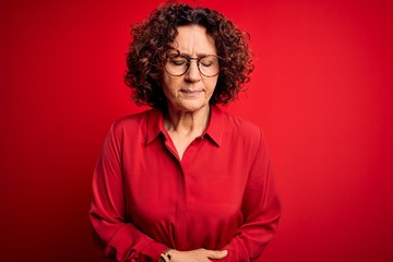 Middle age beautiful curly hair woman wearing casual shirt and glasses over red background with hand on stomach because indigestion, painful illness feeling unwell. Ache concept.