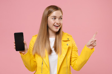 Fototapeta premium Funny young woman girl in yellow leather jacket isolated on pastel pink background. People lifestyle concept. Mock up copy space. Hold mobile phone with blank empty screen point index finger aside up.