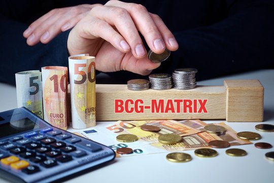 BCG-Matrix. M&auml;nnliche Hand stapelt Geld-Turm (Euro). Begriff an Baustein. M&uuml;nzen, Scheine & Taschenrechner. Konzept f&uuml;r Business, Finanzen, Statistik