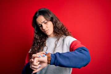 Young beautiful woman with curly hair wearing casual sweatshirt over isolated red background Checking the time on wrist watch, relaxed and confident