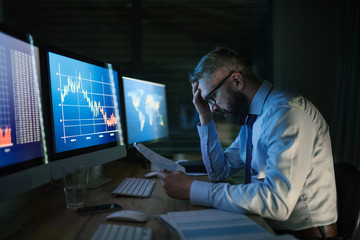 Frustrated businessman with computer sitting at desk, working late. Financial crisis concept.