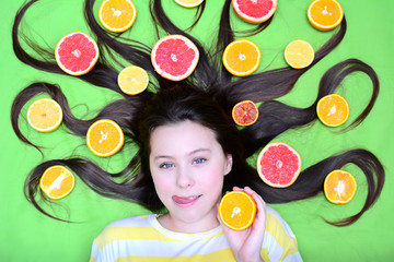 Obraz na płótnie Canvas A cheerful young girl lies with citruses in her loose hair, holds a circle of orange in her hands and licks her lips. Top view