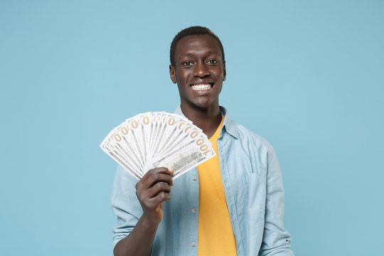 Smiling young african american man guy in casual shirt, yellow t-shirt isolated on blue background in studio. People lifestyle concept. Mock up copy space. Hold fan of cash money in dollar banknotes.