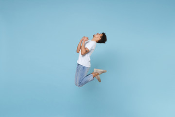 Happy young african american guy in casual white t-shirt posing isolated on pastel blue wall background studio portrait. People lifestyle concept. Mock up copy space. Jumping, doing winner gesture.
