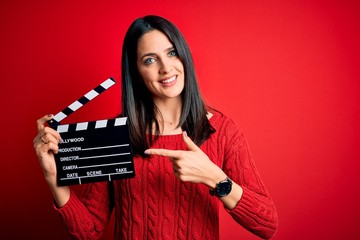 Young director woman with blue eyes making movie holding clapboard over red background very happy...
