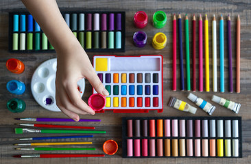 Hand arranging stationery flat lay: pastel crayons, colored pencils, watercolors on wooden background. Art workspace school concept. Top view