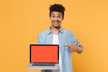 Smiling young african american guy in casual blue shirt isolated on yellow background. People lifestyle concept. Mock up copy space. Point index finger on laptop pc computer with blank empty screen.