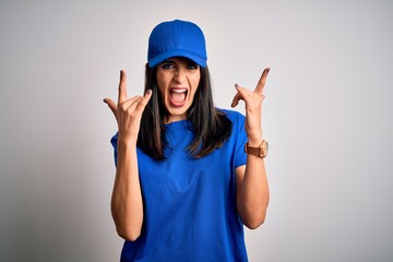 Young delivery woman with blue eyes wearing cap standing over blue background shouting with crazy expression doing rock symbol with hands up. Music star. Heavy concept.