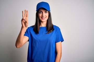 Young delivery woman with blue eyes wearing cap standing over blue background showing and pointing up with fingers number three while smiling confident and happy.