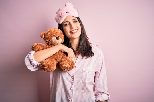 Young brunette woman with blue eyes wearing pajama hugging teddy bear stuffed animal with a happy face standing and smiling with a confident smile showing teeth