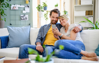 Happy couple in love sitting indoors at home, using smartphone.