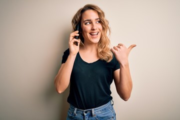 Beautiful blonde woman having conversation talking on the smartphone over white background pointing and showing with thumb up to the side with happy face smiling