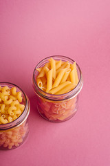 Two glass jars with variety of uncooked golden wheat pasta on minimal pink background, angle view copy space