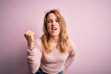 Naklejka na ściany i meble Young beautiful blonde woman wearing casual sweater and sunglasses over pink background angry and mad raising fist frustrated and furious while shouting with anger. Rage and aggressive concept.