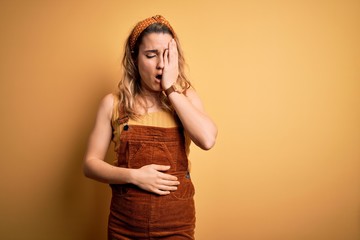 Young beautiful blonde woman wearing overalls and diadem standing over yellow background Yawning tired covering half face, eye and mouth with hand. Face hurts in pain.