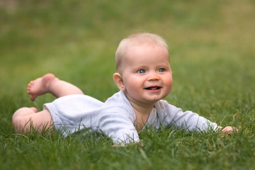 Little baby boy with blue eyes is lying on green grass in the garden and smiling