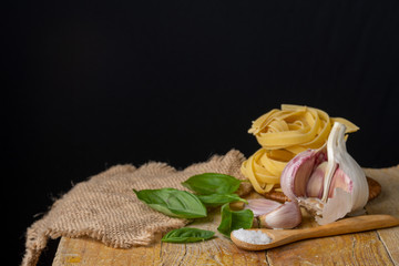 Top view of garlic, basil, tagliatelle, with burlap on wooden table, black background, horizontally, with copy space