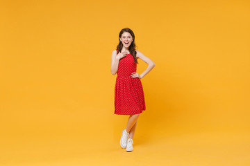 Excited funny young brunette woman girl in red summer dress posing isolated on yellow wall background studio portrait. People sincere emotions lifestyle concept. Mock up copy space. Put hand on chest.