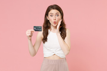 Amazed young brunette woman girl in light casual clothes posing isolated on pastel pink wall background studio. People lifestyle concept. Mock up copy space. Hold credit bank card, put hand on cheek.