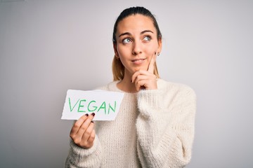 Young beautiful blonde woman with blue eyes holding paper with vegan message serious face thinking...