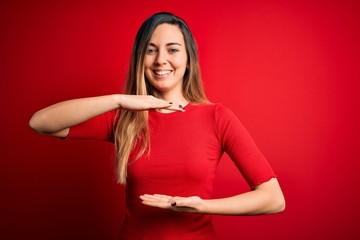 Young beautiful blonde woman with blue eyes wearing casual t-shirt over red background gesturing with hands showing big and large size sign, measure symbol. Smiling looking at the camera. Measuring