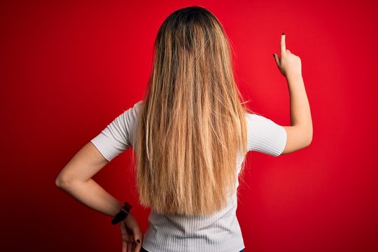 Beautiful blonde woman with blue eyes wearing casual white t-shirt over red background Posing backwards pointing ahead with finger hand