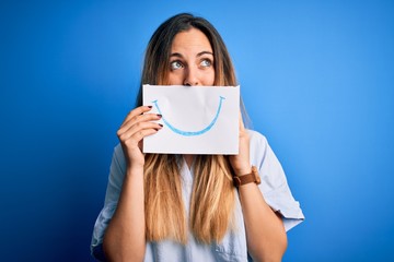 Young beautiful brunette woman holding card with smile illustration on mouth serious face thinking...