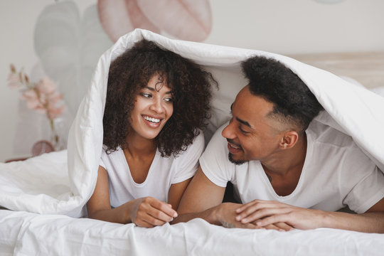 Young calm african loving couple girl guy in t-shirts lying on bed with white sheet pillow blanket indoors in bedroom at home, spending time in room. Rest relax good mood quarantine lifestyle concept.
