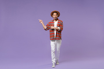 Smiling young african american guy in casual colorful shirt hat posing isolated on violet wall background studio portrait. People lifestyle concept. Mock up copy space. Pointing index fingers aside.