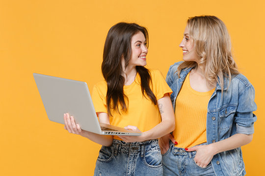 Two smiling young women girls friends in casual t-shirts denim clothes isolated on yellow background. People lifestyle concept. Mock up copy space. Working on laptop pc computer looking at each other.