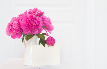 Pink peonies in white enamelled vase. Beautiful flowers in interior design. White paper for invitation text, white peonies in a vase, interior decoration.