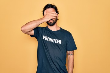 Young handsome hispanic volunteer man wearing volunteering t-shirt as social care covering eyes...