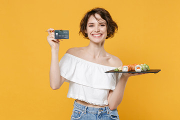 Young woman girl in casual clothes hold in hand credit card makizushi sushi roll served on black plate traditional japanese food isolated on yellow background studio portrait. People lifestyle concept
