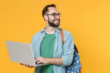 Smiling young man student in casual clothes glasses with backpack isolated on yellow wall background studio. Education in high school university college concept. Hold laptop pc computer looking aside.