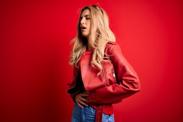 Fototapeta premium Young beautiful blonde woman wearing casual jacket standing over isolated red background with hand on stomach because nausea, painful disease feeling unwell. Ache concept.