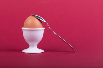 one brown egg in white egg cup before red background, a teaspoon is laying on it