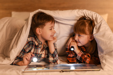 Little children reading book under blanket at night