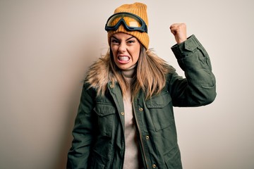 Young brunette skier woman wearing snow clothes and ski goggles over white background angry and mad raising fist frustrated and furious while shouting with anger. Rage and aggressive concept.