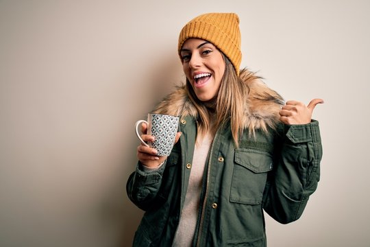 Young beautiful brunette woman wearing snow clothes drinking mug of coffee pointing and showing with thumb up to the side with happy face smiling