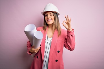 Young beautiful brunette architect woman wearing safety helmet holding blueprints doing ok sign...