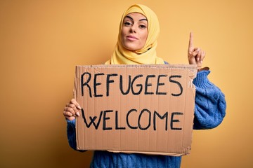 Beautiful arab woman wearing islamic hijab holding banner with refugees welcome message surprised...