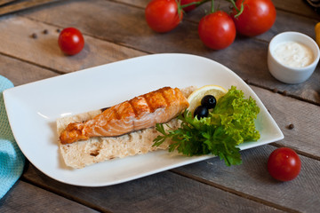 Grilled salmon steak served on a white plate with lemon and herbs. wooden background