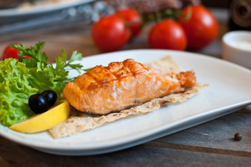 Grilled salmon steak served on a white plate with lemon and herbs. wooden background
