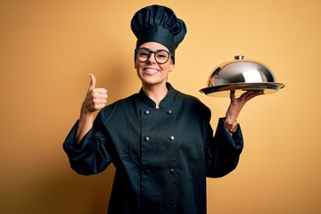 Young beautiful brunette chefwoman wearing cooker uniform and hat holding tray with dome happy with big smile doing ok sign, thumb up with fingers, excellent sign