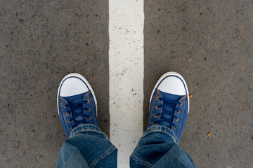 Male feet in blue jeans and blue sneakers shoes stand on street asphalt pavement with dividing line