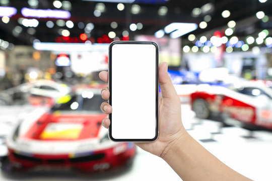 Mock up blank white screen mobile phone for automotive advertisement. Hand holding smartphone with Blurred image of super cars and sport cars in exhibition hall. Mockup for your artwork.