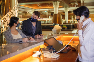 Couple and receptionist at counter in hotel wearing medical masks as precaution against virus. Couple on a business trip doing check-in at the hotel