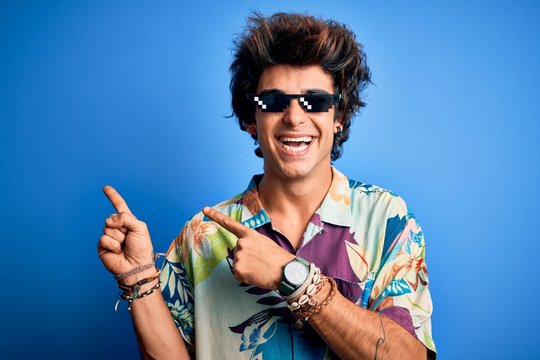 Young handsome man wearing thug life glasses standing over isolated blue background smiling and looking at the camera pointing with two hands and fingers to the side.