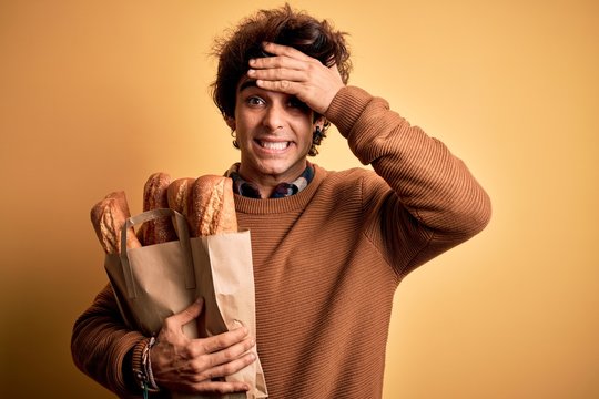 Young handsome man holding paper bag with bread standing over isolated yellow background stressed with hand on head, shocked with shame and surprise face, angry and frustrated. Fear and upset